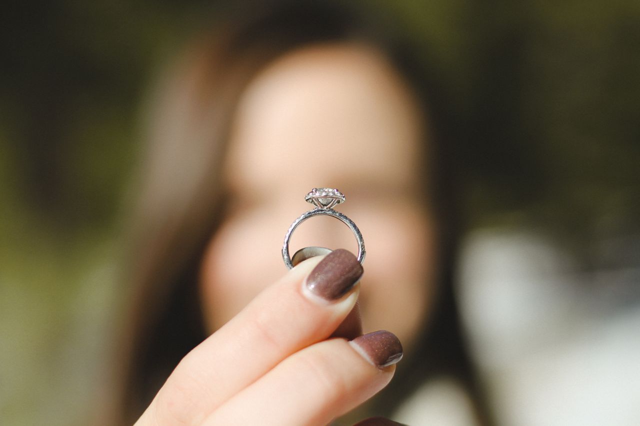 Engagement ring with intricate metalwork details seen from the side profile. The ring is held by a blurry figure in the background Engagement ring with intricate metalwork details seen from the side profile. The ring is held by a blurry figure in the background
